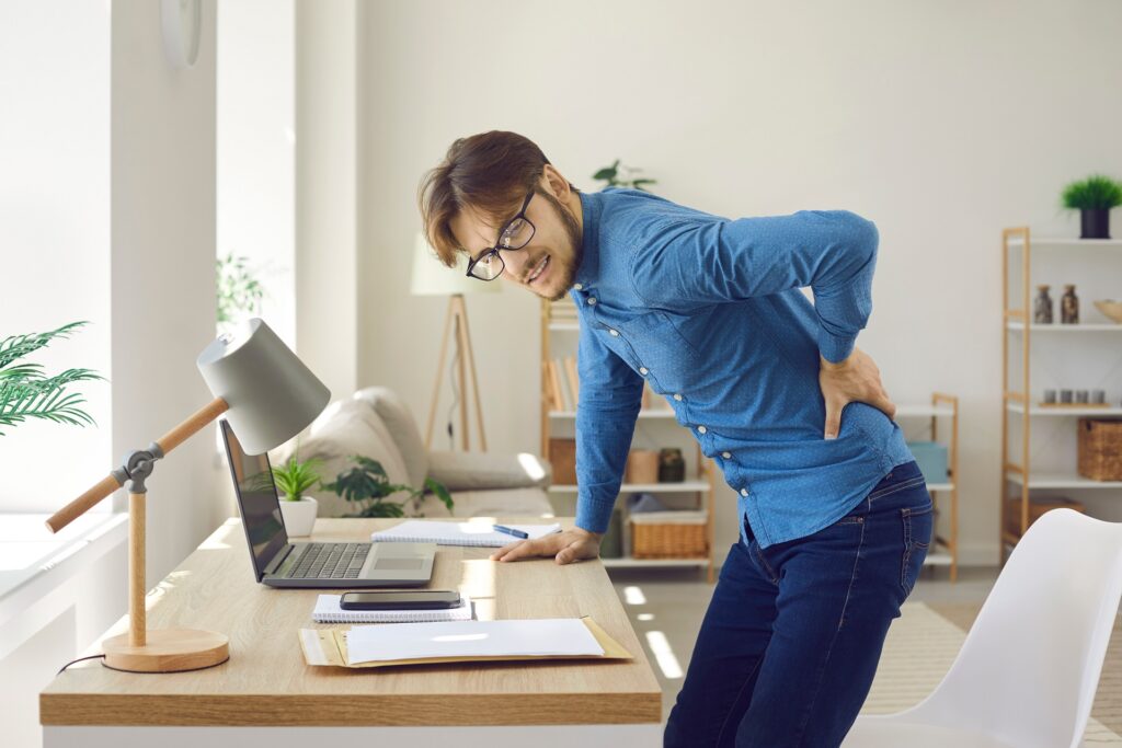 Male office worker experiences severe back pain caused by prolonged sitting in wrong position. Unhappy man experiencing spasm of spine getting up from behind workplace in office.Sedentary life concept | Hudson Premier PT & Sports | Jersey City | Union City | NJ